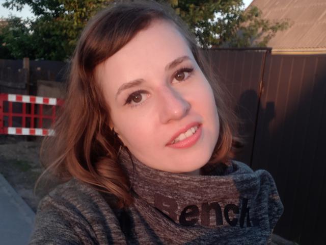 RosaJillian has fair skin, brown eyes, and shoulder-length brown hair. She's wearing a gray turtleneck, smiling with pink lips, and the image is a daytime selfie.