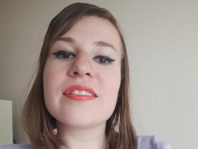 RosaJillian has fair skin, brown eyes, black eyeliner, red glossy lips, and straight brown hair. Her face is centered, smiling, with a slight upshot. She's wearing a purple top.