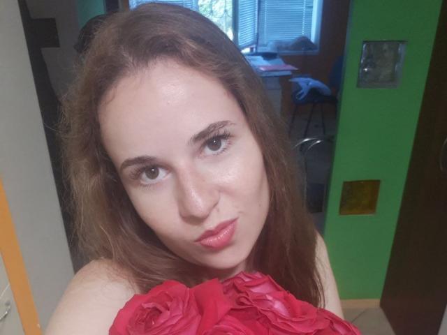 RosaJillian with wavy brown hair, light skin, and red lipstick, wearing a red floral top, holding it up to her tits, in a green-walled room, looking at camera.