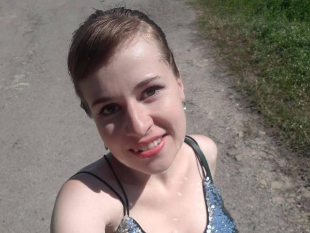 RosaJillian with short brown hair, fair skin, small tits, wearing a sparkly blue top, smiling with red lipstick, standing on a gravel path with green grass.