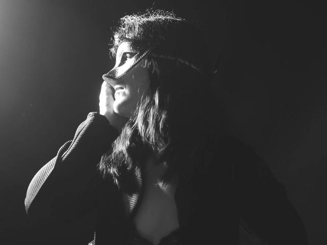 Black-and-white photo of sweetSilvana-hot with wavy hair, wearing a mask and dark sweater, illuminated from the left, showing her profile with a gentle smile.
