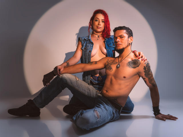 AbbyAndVincent with red hair, small tits, and no bra, sits on the floor wearing a denim vest and jeans. Vincent, shirtless with tattoos, sits beside her, touching her.