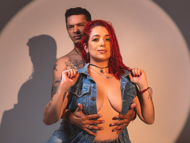 AbbyAndVincent, con cabello rojo, senos grandes, vistiendo un chaleco vaquero abierto, siendo sostenida por Vincent desde atrás, quien tiene tatuajes y está sin camisa, con las manos sobre sus senos..