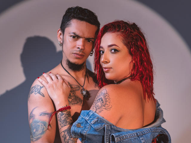 AbbyAndVincent, with red hair, looking over shoulder, topless, denim jacket off, tattoos, holding shirtless Vincent, tattoos, nose ring, shadow, intense gaze, arm bracelet, warm light.