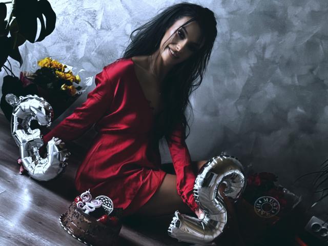KinkyAmelie con lunghi capelli neri, vestito rosso, seduta sul pavimento, mani sui palloncini metallici a forma del numero "6", pavimento di legno, fiori sullo sfondo, sorridente, sexy, carina, pelle chiara..