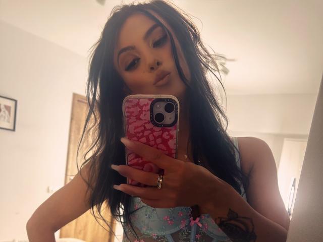 KinkyAmelie with long black hair, wearing a light blue floral top, taking a mirror selfie with a pink cracked phone, showing a nose ring and ring on her right hand.