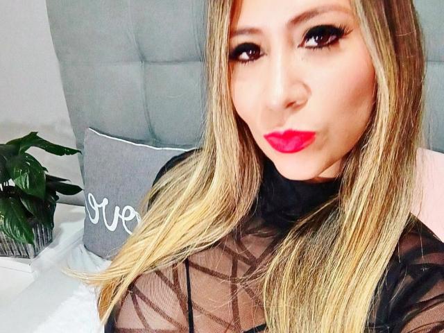 Camilover with blonde hair, black sheer top, red lipstick, and heavy eyeliner, pouting slightly, showing off her tits. She's on a bed with a grey headboard.
