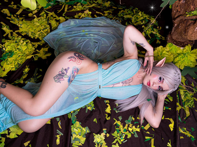 MelanyBlair with pale skin, long silver hair, elf ears, and tattoos, lying in a blue sheer dress, showing her ass and tattoos, on yellow and green leafy fabric.
