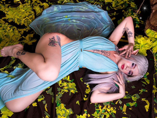 MelanyBlair with pale skin, platinum blonde hair, blue dress, lying on black and yellow leaf-covered ground, showing tattoos, her ass, and her hand near her pussy.