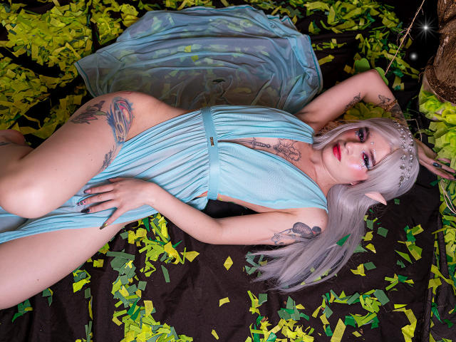 MelanyBlair lies on a black and green leaf-covered surface, wearing a blue, sheer dress, exposing her tits and pussy. She has silver hair, elf ears, and multiple tattoos.