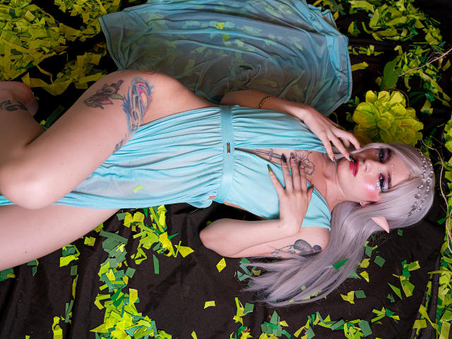 MelanyBlair with long silver hair, elf ears, and tattoos, lying in a sheer blue dress, touching her red-lipsticked mouth, surrounded by yellow green leaves on a black surface.