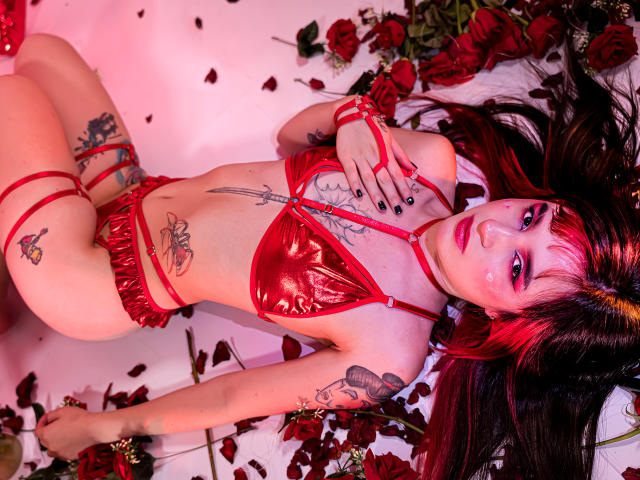 MelanyBlair with red metallic bikini, tattoos, and red rose petals, lying on white floor, red and pink lighting, black nail polish, long dark red hair, looking up.