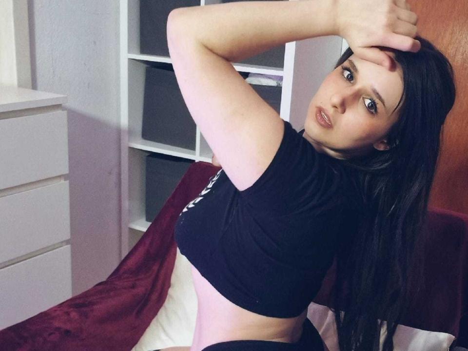 JenniSexy with long brown hair, black crop top, and black bottoms, looking at camera with arm raised, showing her tits, sitting on bed with red blanket.