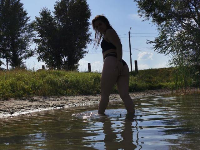 VioSweet stands in shallow water, wearing a black bikini, showing her ass, with wet skin and long brown hair, looking back over her shoulder.