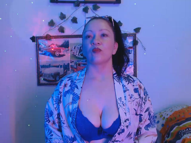 CharloteEzra with big tits, wearing a blue bra and floral robe, has dark hair in a ponytail, blue and pink lighting, and a subtle smile. Background with wall decor.