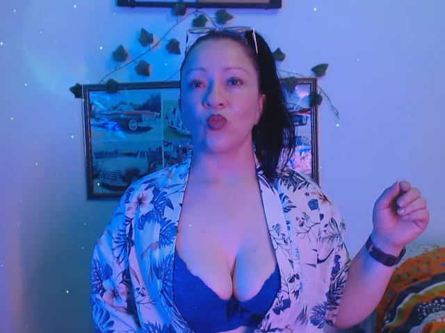 CharloteEzra with blue-lit skin, pouty lips, and big tits in a floral shirt, open to reveal a blue bra. She's wearing a watch and has her hair in a ponytail.