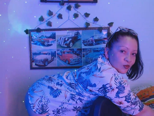CharloteEzra with blue floral dress, bending over, looking back, medium tits, dark hair, red lipstick, wall with car photos, string lights, and green leaves above.