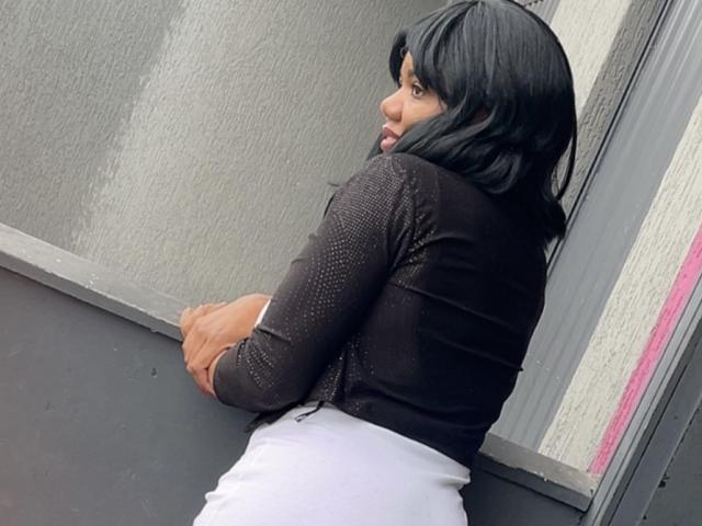 BarbaPapaz with black hair, wearing a black, long-sleeve, sparkly top and white pants, leaning against a grey wall, looking over her shoulder with a slight smile.
