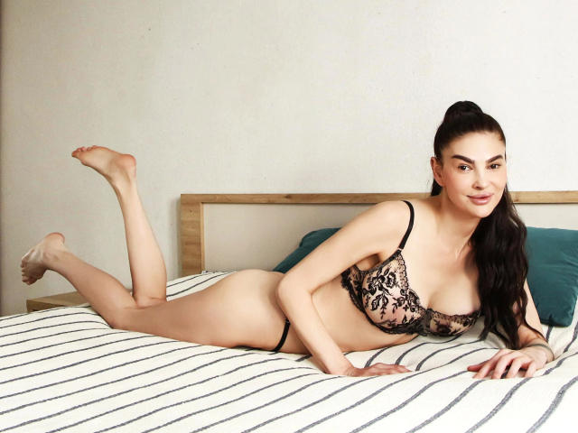 ReifeSuzanna-hot with long dark hair, lying on a bed in black lace lingerie, showing her tits and ass, legs up, smiling at the camera.