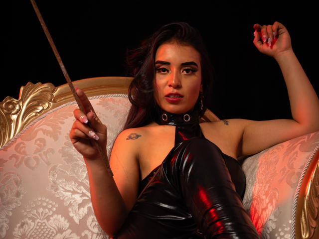 MelizaQueen with dark hair, wearing black latex, holding a whip, sitting on an ornate chair, revealing tattoos, red nails, and a confident expression.