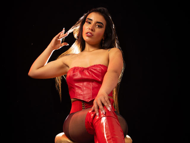 MelizaQueen with long dark hair, red leather corset, red high boots, and sheer black pantyhose, sitting, hand in hair, black background, looking seductively at camera. Boobs, ass, pussy.
