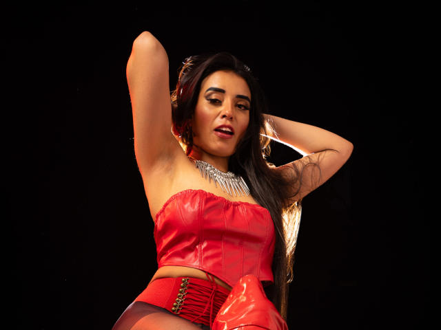 MelizaQueen con una giacca di pelle rossa, che mostra il seno, braccia alzate, lunghi capelli neri, collana scintillante, sfondo nero, aspetto sexy, sicuro di sé e leggermente a bocca aperta..