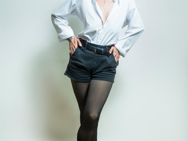 AriiannaHot with a white button-up shirt, black shorts, and black sheer pantyhose, hands on hips, showing her tits slightly, standing against a white wall.