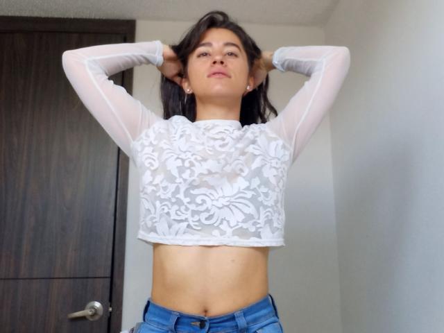 MaddieHottie with hands in her dark hair, wearing a white sheer lace crop top, showing her tits, and blue jeans, standing in a room with a wooden door.