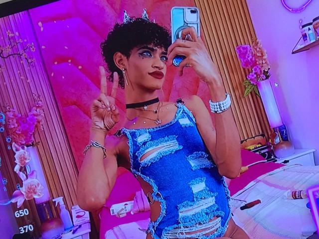 AdharaX69 with curly black hair, blue ripped dress, black choker, and silver jewelry, making a peace sign, holding a phone, pink and purple room, wearing a cat-eye makeup.
