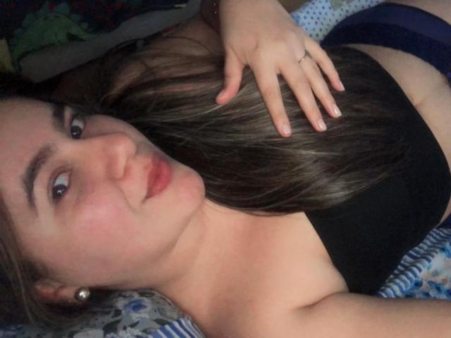 EmaSmit with long brown hair, black tube top, and blue panties, lying on bed, hand in hair, pouty lips, earrings, and a ring on her finger.