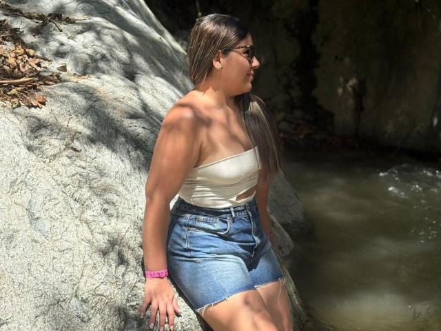 EmaSmit with long brown hair, wearing a white strapless top and denim shorts, sitting on a rock by a stream, sunlight filtering through leaves. Her ass is slightly visible.
