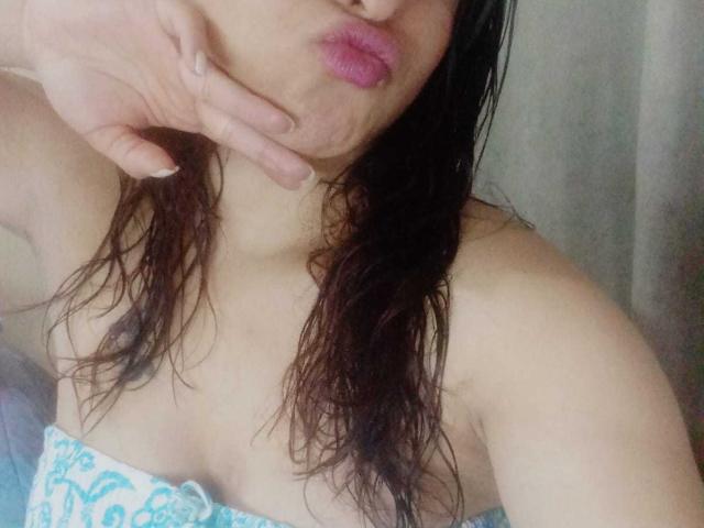 AntonellaWayne's pouty pink lips, wet brown hair, and cleavage in a blue and white patterned top, with her hand near her mouth, showing off her tits.