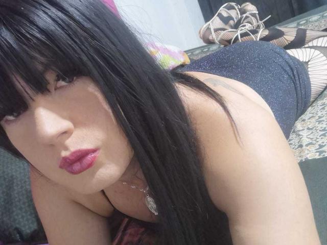 BonnySmithTs with long black hair, purple lipstick, and a silver necklace, lying on a bed, wearing a sparkly black dress, showing her ass, and striped heels in the background.