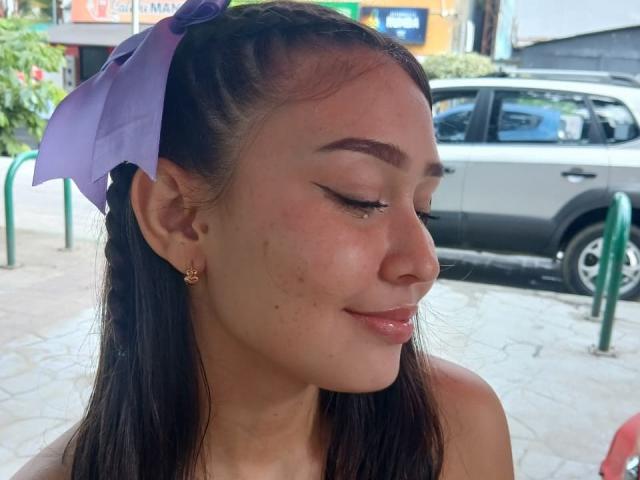 BellaMia, met een paars lint in haar donkere haar, gesloten ogen, subtiele make-up en een klein sproetje op haar wang, glimlachend, met gouden oorbellen, op een foto..