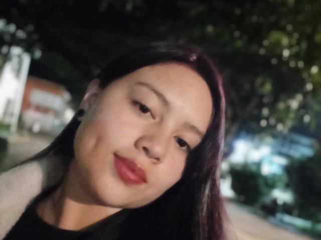 Il viso di NatalliieSweet, leggermente inclinato, con capelli neri e lisci, pelle chiara, rossetto rosso, una maglietta nera e auricolari neri. Notte, sfondo sfocato, illuminazione soffusa..