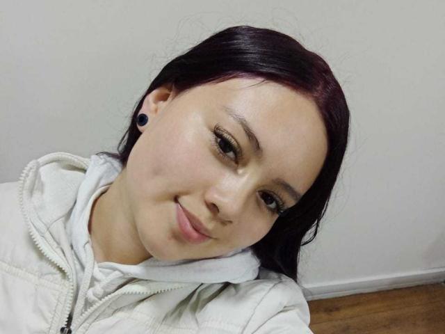NatalliieSweet with dark red hair, black ear gauges, white hoodie, and a slight smile, looking at the camera. She has light skin and a cute, relaxed face.