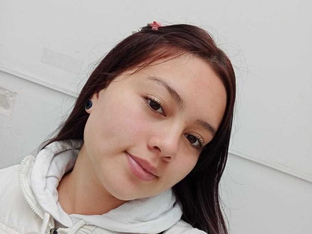 NatalliieSweet, con occhi marroni chiari, pelle chiara, piccole orecchini neri, e capelli castano scuro, indossa una felpa bianca, sorride dolcemente e guarda la fotocamera..