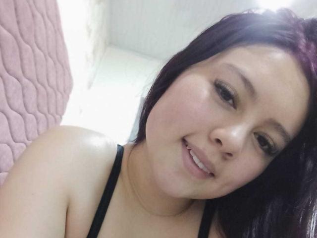 NatalliieSweet, con cabello oscuro, sonriendo, vestida con un top negro de tirantes, tiene una piel lisa, pecho superior visible, y está ligeramente inclinada, con luz en su rostro..