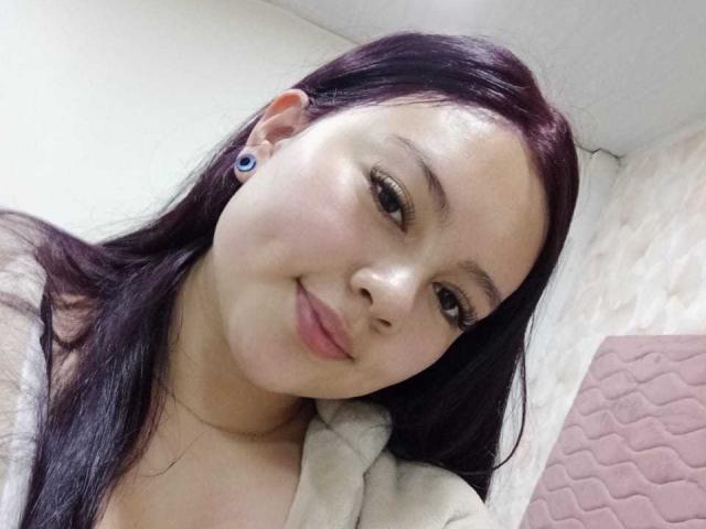 NatalliieSweet, con i capelli viola scuro, i piercing alle orecchie blu e un sorriso appena accennato, indossa una maglia beige, mostrando il suo viso liscio e chiaro e le sue piccole labbra rosa..