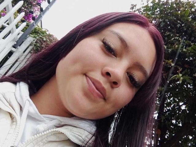 NatalliieSweet mit dunkelviolettem Haar, langen Wimpern und einem leichten Lächeln, trägt eine weiße Kapuzenjacke und blickt nach unten in die Kamera. Ihre Nase ist klein und ihre Haut ist hell..