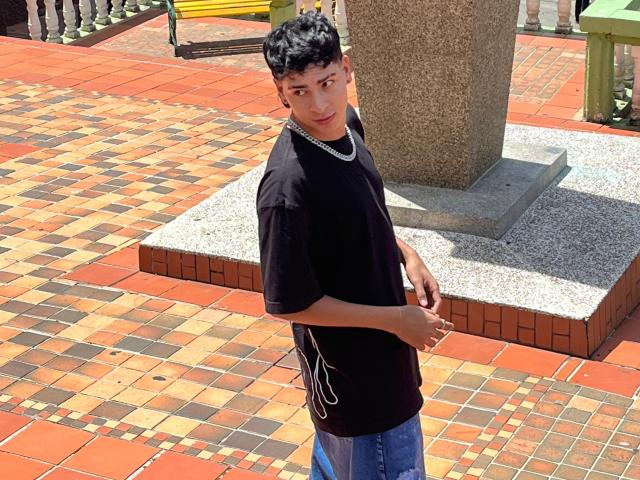 Adoniss mit lockigem, schwarzem Haar, der ein schwarzes T-Shirt und blaue Jeans trägt, eine silberne Kette um den Hals und weiße Bänder, steht auf einem mit Ziegeln gemusterten Boden und blickt zurück..