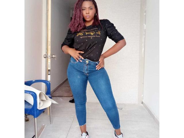 MacarenaBoobs has dark skin, red curly hair, wears a black polka-dot shirt, blue high-waisted jeans, and white sneakers, standing with hands on hips, looking at camera.