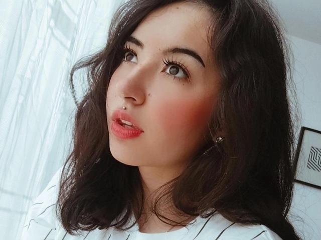 SusanMonroy has wavy dark brown hair, a nose piercing, red blushed cheeks, and full pink lips, wearing a white striped shirt, with a subtle smile, looking slightly up.