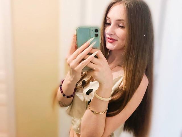 PamelaXu with long brown hair, smiling, holding a green phone, wearing a beige top, showing her tits, light makeup, pink lipstick, purple bracelet, and gold bracelet in a mirror selfie.