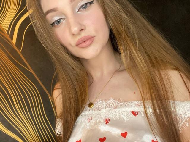 PamelaXu has long brown hair, blue eyes with winged eyeliner, pink lips, and wears a white lace-off-shoulder top with red heart patterns and a gold heart necklace.