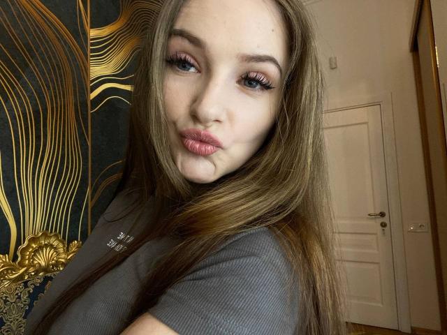 PamelaXu has long brown hair, full pink lips, and heavy eyelashes. She's wearing a gray ribbed top, showing her tits slightly. Her expression is pouty.