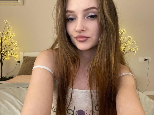 PamelaXu with long brown hair, pale skin, and pink lips, wearing a white tank top, showing her tits slightly, looking into the camera with a neutral expression.