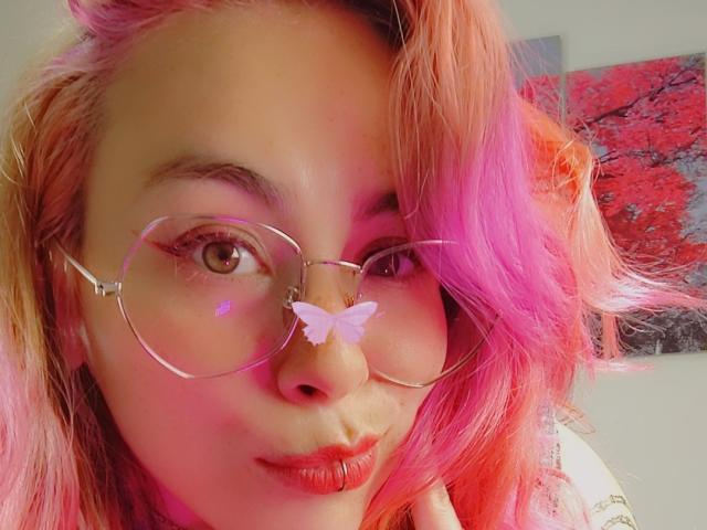 NatashaLau with pink and orange hair, wearing clear glasses with a pink butterfly sticker, puckering red lips, brown eyes, soft smile, and fair skin.