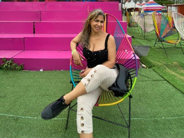 BrigiteWalsh, with tan skin and long brown hair, sits on a colorful chair, wearing a black top, white ripped pants, and black shoes, smiling at the camera.