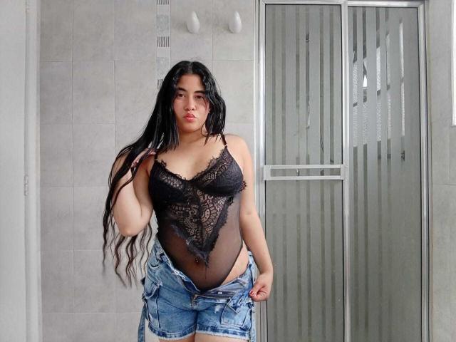 BiancaSolas mit langen, schwarzen Haaren, die schwarze Spitzendessous, die ihre Brüste zeigen, und ungeknöpfte blaue Jeans trägt, steht in einem Badezimmer vor einer Glastür zur Dusche..